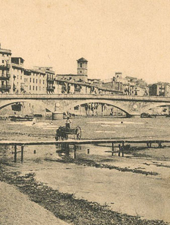 Vista del riu Onyar al seu pas pel centre de la ciutat de Girona. En primer terme, un grup de dones rentant al riu, al costat de la palanca de l'Areny. A la dreta part de la muralla del riu Onyar i el portal de l'Àngel. En segon terme, el pont de Pedra. Al fons sobresurt el campanar de l'església de les Bernardes. 1900-1903
