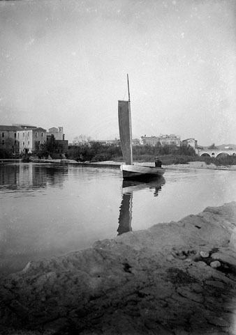 Francisco Mitjans dins una barca amb una vela navegant per la resclosa de Sarrià de Ter. Al fons a la dreta s'observa el pont de l'Aigua, i a l'esquerra l'església de Sarrià de Ter. 1917-1918
