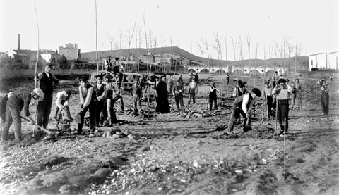 Alumnes de la granja-Escola de Sant Isidre netejant les terres de cultiu de l'escola després d'un aiguat, al barri de Pont Major. Al fons, d'esquerra a dreta, l'església parroquial de Sarrià de Ter i el pont de l'Aigua sobre el riu Ter. 1920-1930