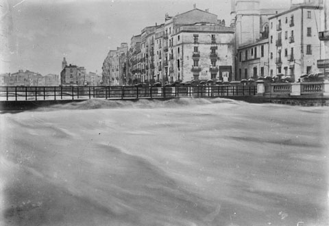 vista del riu Onyar durant l'aiguat de l'any quaranta on apareix el pont del Pes de la Palla superat per l'aigua. A la dreta s'observa la rambla Verdaguer i algunes persones amb paraigües contemplant el riu. 1940