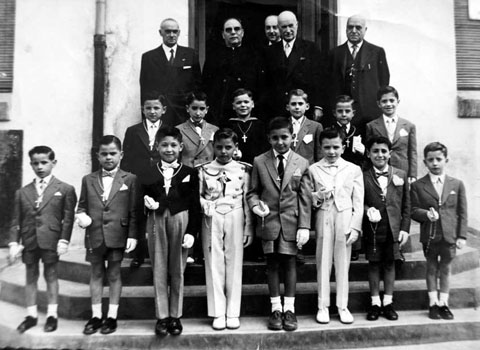 Retrat d'un grup de nens de les Escoles d'Ensenyament Primari de Foment de Cultura vestits de Primera Comunió. Els acompanya, entre d'altres, mossèn Joaquim Batlle. Ca. 1960