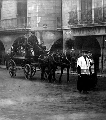 Seguici fúnebre d'un enterrament al seu pas per la plaça del Vi, amb el carruatge tirat per cavalls engalanats. A la dreta, façana del Banc Espanyol de Crèdit. 1920