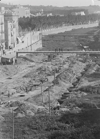 Obres al llit del riu Onyar per a la construcció del Mercat Municipal projectat per Maggioni. S'observen el pont del Pes de la Palla i el del Carme. A l'esquerra, una parada d'autobusos i, en el segon pis del primer edifici, un cartell que anuncia la ubicació del Tribunal Tutelar de Menors. 1929