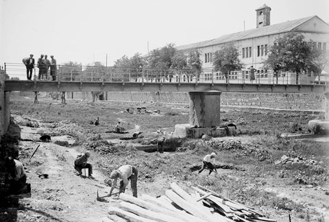 Obres al llit del riu Onyar per a la construcció del Mercat Municipal projectat per Maggioni. Algunes persones s'ho miren des del pont del Pes de la Palla. A la part superior s'observa el convent de Sant Francesc de Paula, conegut popularment com els Mínims, en funcions de caserna i magatzem militar. 1929