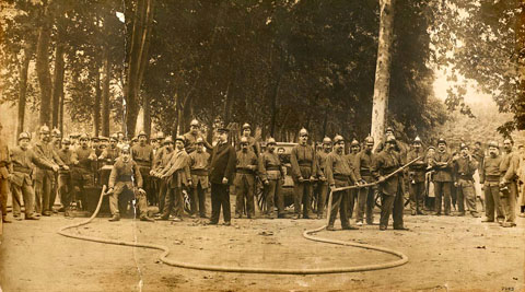 Retrat de grup de la Primera Companyia de Bombers de l'Ajuntament de Girona, amb els seus uniformes i recursos materials per lluitar contra el foc, a la Devesa. Possiblement es tracti d'una de les exhibicions que es portaven a terme durant les fires. 1900