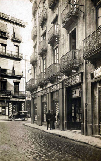 El carrer de la Cort Reial i la plaça de l'Oli. En primer terme, l'establiment de Fotografia Lux i la Llibreria Escolar. Al fons, la farmàcia Roca. 1930-1940