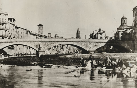 Dones rentant la roba al riu Onyar, davant del pont de Pedra. A l'esquerra, el campanar de les Bernardes i al seu costat, el del convent de Santa Clara. Al centre, entre els arcs del pont de Pedra, s'entreveu l'antiga palanca de les Peixateries Velles que seria substituïda pel pont construït per Eiffel. 1870