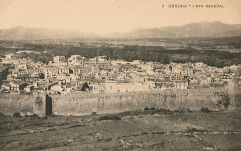 Vista del barri del Mercadal des de la muntanya de les Pedreres. En primer terme el tram de la muralla comprés entre la torre del Portal Nou i la torre dels Socors. Al fons, d'esquerra a dreta, les torres de l'església del Sagrat Cor, les naus de la fàbrica Grber i el campanar de l'església de Santa Susanna i de les Bernardes. Al centre, la plaça de Braus de Girona. 1910-1918